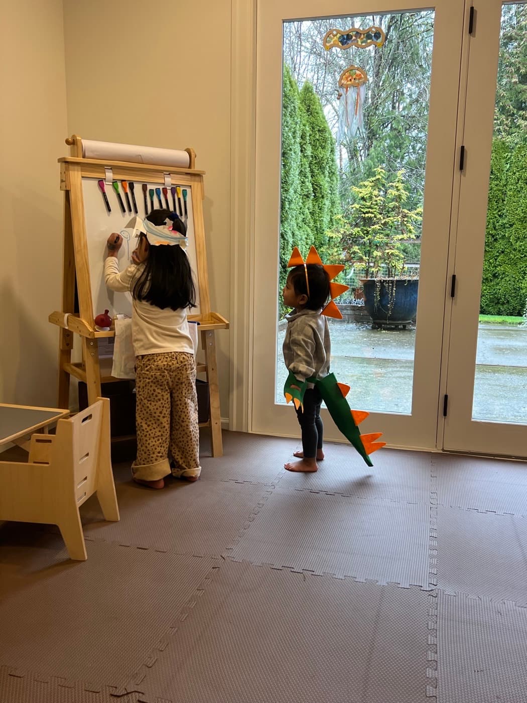 Our daughters mid-lesson — one drawing at the easel, one in a homemade dinosaur costume watching the rain
